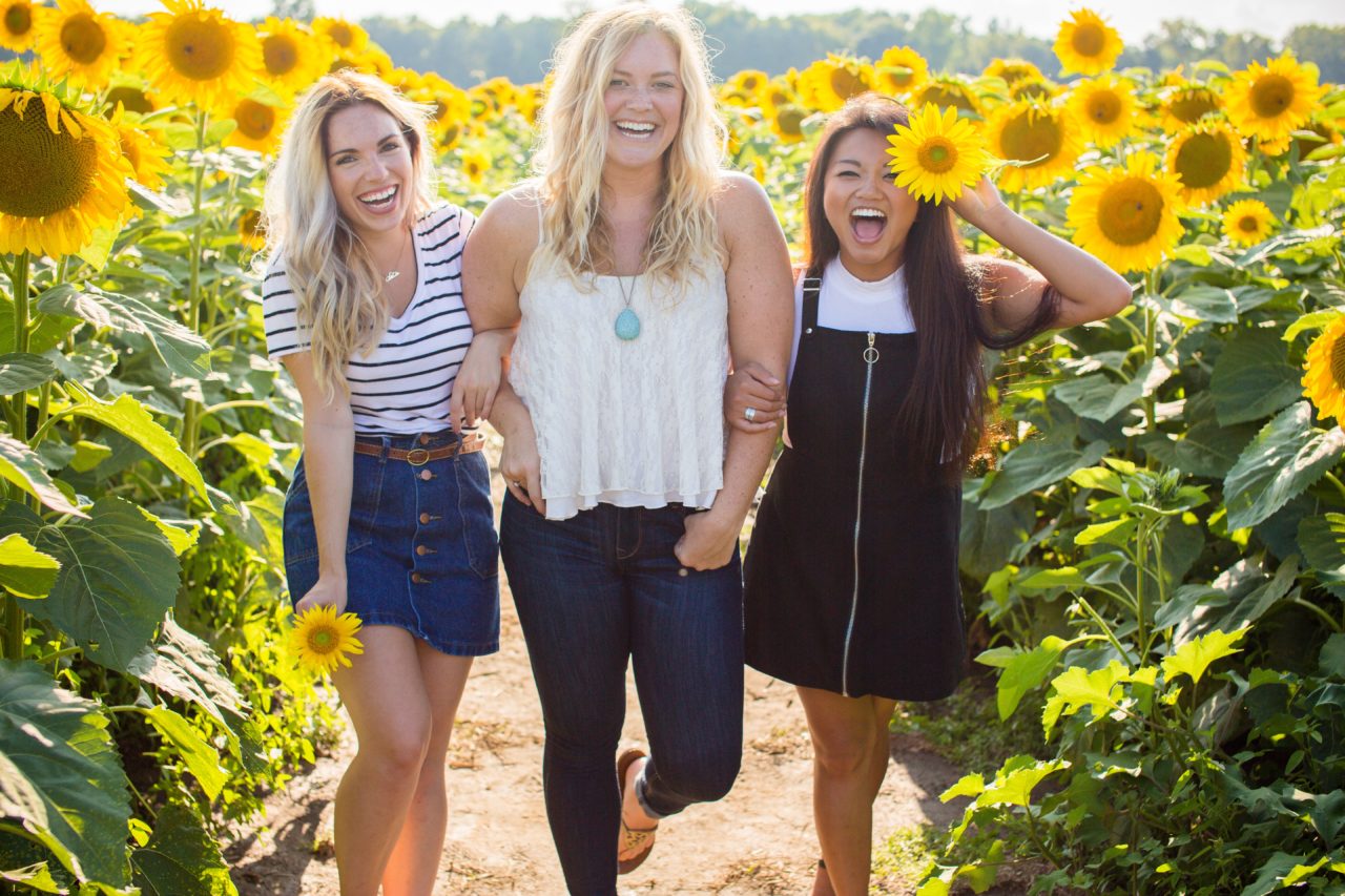three women with glowing skin in a sunflower field laughing