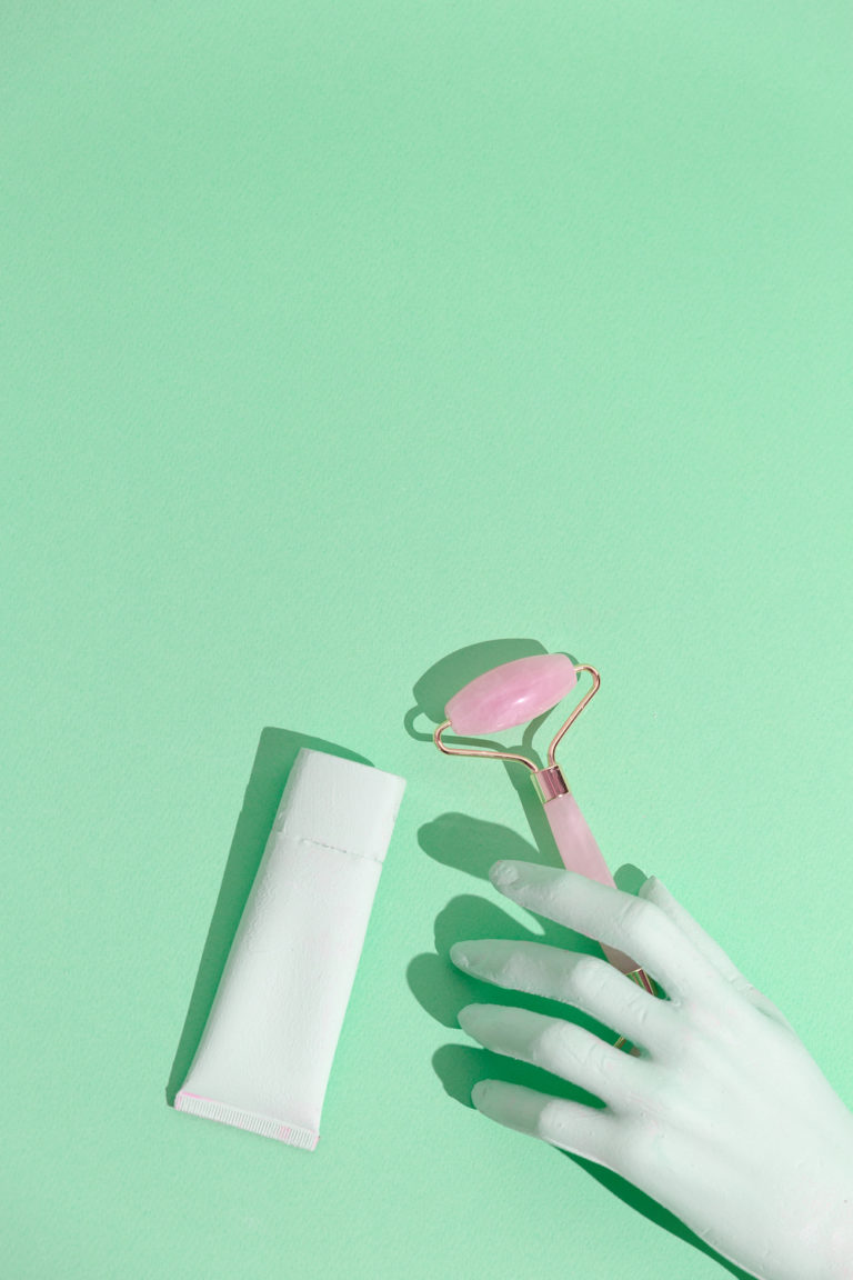 pink skincare roller and white bottle on a green background with white gloves