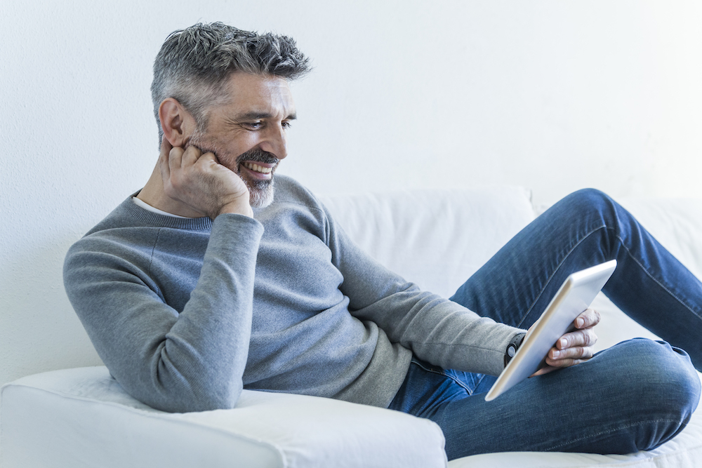mature man smiling and looking at a tablet on a white couch
